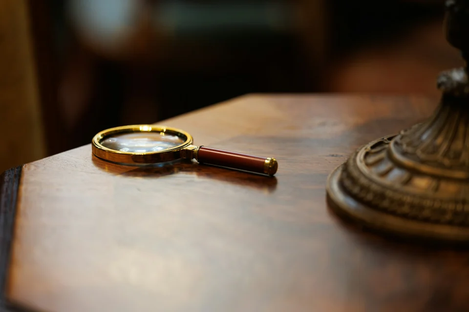 Brass-rimmed magnifying glass on a polished mahogany table, with an ornate bronze lamp base at right