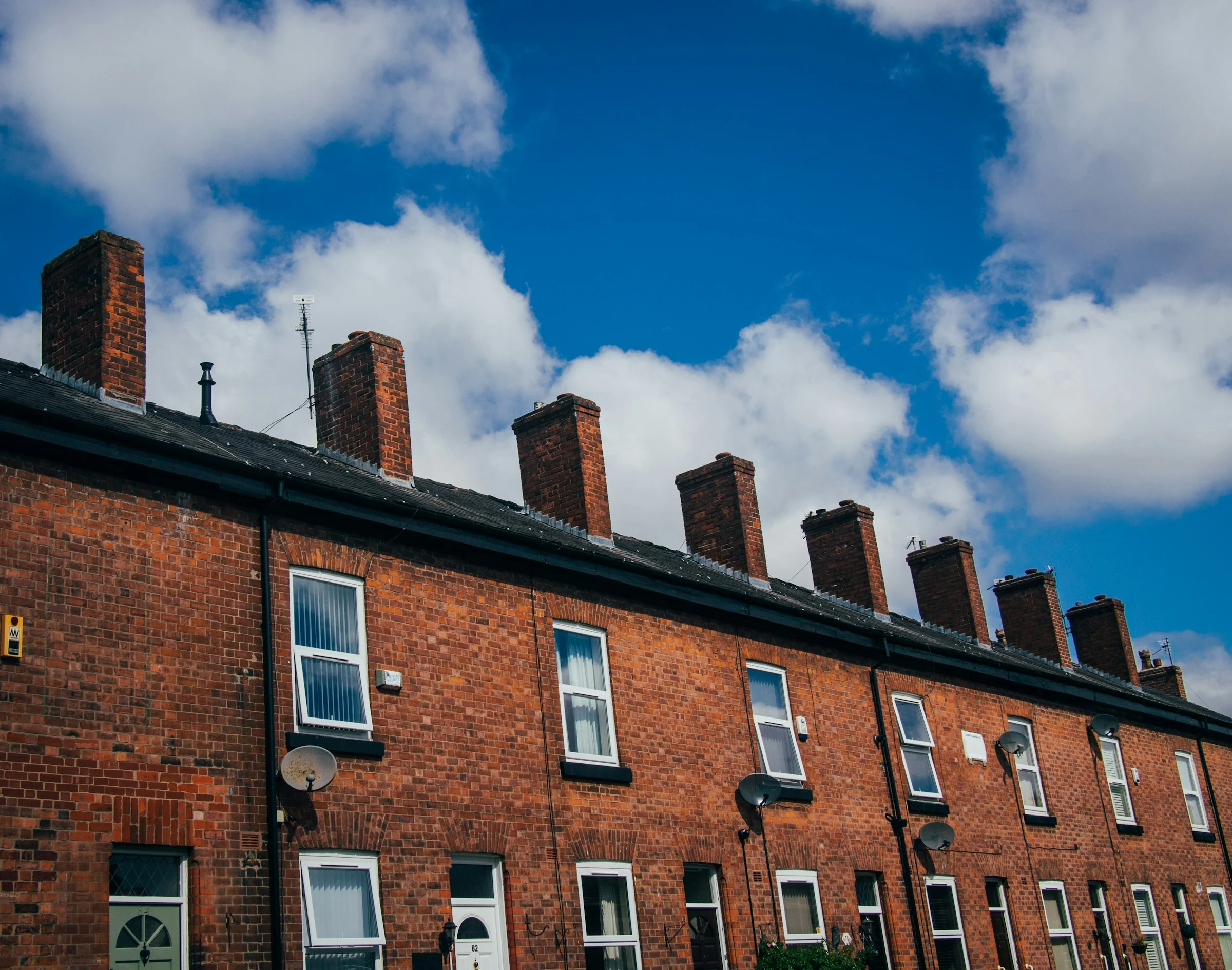 Row of red-brick terraced houses with chimney stacks and satellite dishes under a blue sky