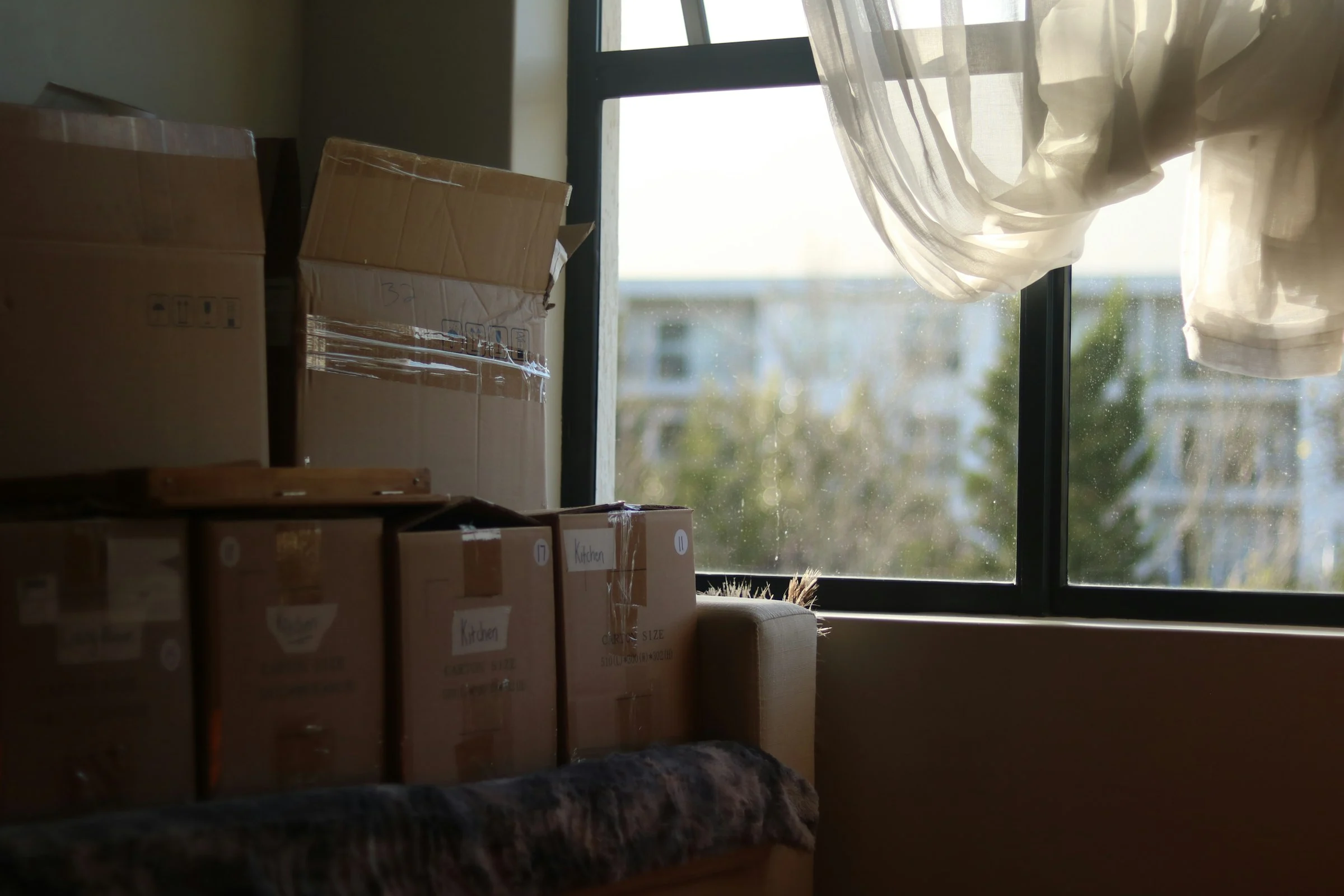 Taped boxes marked Kitchen stacked on a sofa beside a window with a sheer curtain